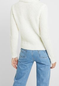 White knitted sweater with a high collar, ribbed texture, and fitted cuffs, paired with light blue wide-leg jeans.
