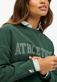 Sudadera verde con "ATHLETICO" en texto gris; superpuesta con una camisa de cuello a rayas azules y blancas. El puño presenta un acento blanco.