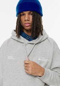 Ljusgrå hoodie med en framficka, tryckt text och dragsko i huvan. Bärs över en denimskjorta och är accessoriserad med en blå stickad mössa.