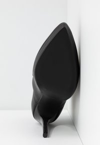 Chaussure à talon haut noire à bout pointu reposant sur le côté contre une surface blanche en angle.