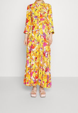 Długa żółta sukienka maxi w kwiaty z długimi rękawami i zapięciem na guziki z przodu, zestawiona z bladoróżowymi sandałami na platformie z krzyżującymi się paskami.