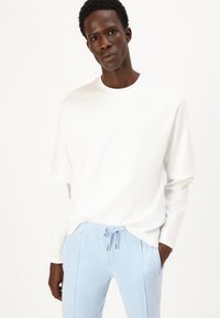 Camicia bianca a maniche lunghe con scollatura rotonda, abbinata a pantaloni azzurri concordi con cordino e dettagli in piping laterali.