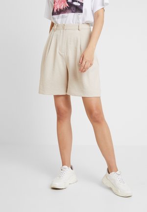 Frau trägt cremefarbene, hoch taillierte, plissierte Shorts mit Knopf und Reißverschluss, weißes Grafik-T-Shirt und weiße klobige Sneaker.