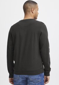 Sudadera verde oscura con escote redondo, puños y dobladillo acanalados. Lleva puesta sobre un vaquero azul, mostrando una textura suave y un diseño ajustado.