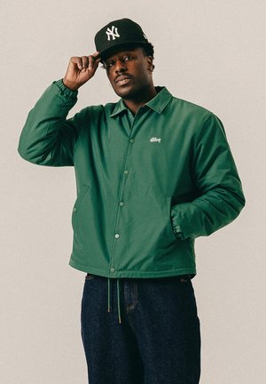 Mand iført grøn knap-op jakke og mørke jeans, justerer sort New York Yankees baseballkasket, står mod en ensfarvet baggrund.