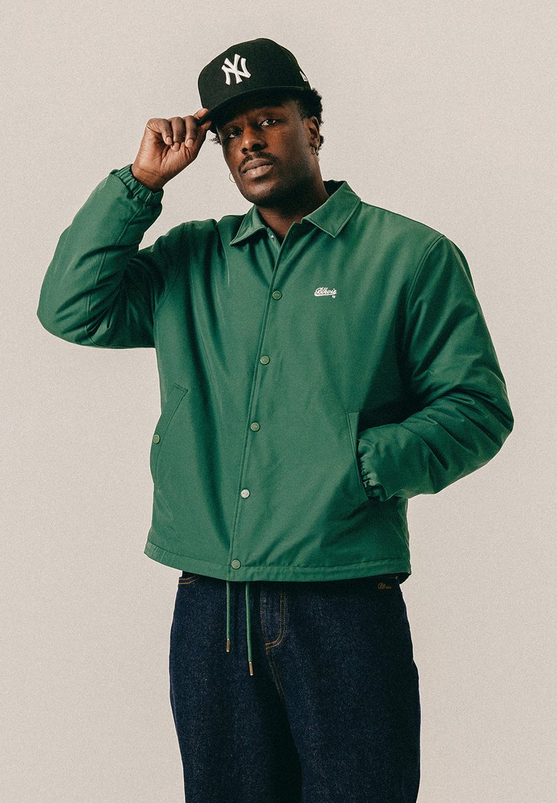 Homme portant une veste verte à boutons et un jean foncé, ajustant une casquette de baseball noire des New York Yankees, debout devant un fond uni.