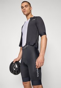 Svart sykkeltrøye med korte ermer og mesh-frontpanel, kombinert med svarte polstrede shorts. Sykkelrytteren holder en svart hjelm.