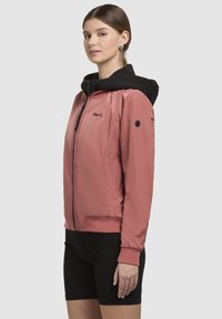 Veste zippée rose clair avec capuche noire, dotée de poignets et d'une taille côtelés, et d'une texture lisse et brillante. Logo sur la poitrine.