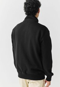Mann mit lockigem Haar, der einen schwarzen Sweatshirt mit hohem Kragen und beigen Hosen trägt, mit dem Rücken zu einer hellen, einfarbigen Hintergrund.