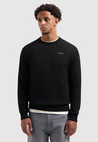 Zwarte sweatshirt van een katoenmix met een ronde hals en geribde manchetten. Kleine logo-accent op de borst. Gecombineerd met lichtgrijze spijkerbroek.