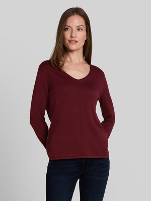 Femme debout, les mains dans le dos, portant un pull bordeaux à manches longues avec col en V et un jean bleu foncé sur un fond gris uni.