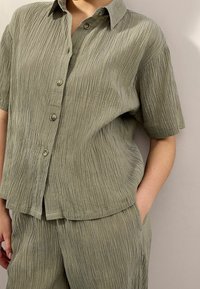 Olive green, short-sleeved, button-up shirt with a textured, waved pattern. Fabric appears lightweight and slightly wrinkled.