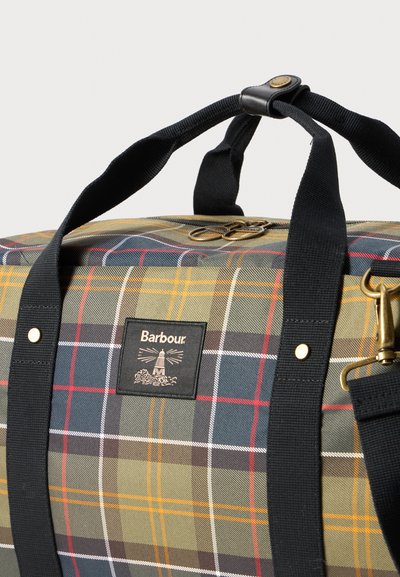 Le sac de voyage présente un motif à carreaux vert, bleu marine et jaune, des poignées noires avec des accents en cuir, et un patch logo sur le côté.