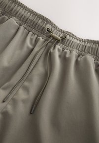 Shorts verts olive avec une texture lisse, taille élastique et cordon de serrage. Présente un design minimaliste et une brillance subtile.