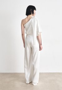 White satin jumpsuit featuring a one-shoulder design with a flowing sleeve and wide-leg fit. Smooth texture with minimalist detailing.
