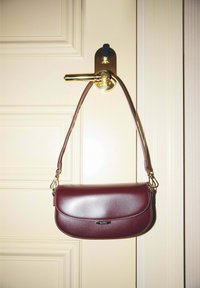 Sac à bandoulière en cuir rouge foncé accroché à une poignée de porte dorée brillante fixée à une porte à panneaux couleur crème.
