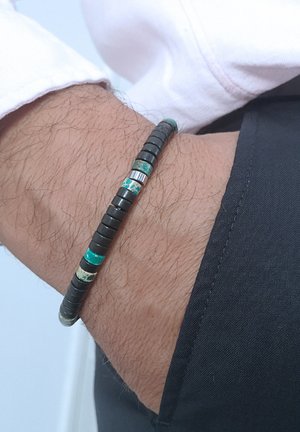 Bracelet en perles noires et turquoises présentant des perles rondes et cylindriques, avec des accents métalliques, porté sur un poignet avec des cheveux clairs.