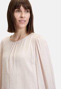 Blusa rosa pallido con scollatura rotonda, maniche arricciate e un sottile dettaglio della tasca. Tessuto morbido e leggero con una texture liscia.