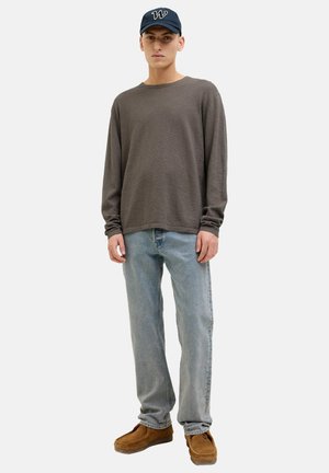 Ung mand iført navy kasket, løs brun langærmet skjorte, lyseblå jeans og brune ruskindssko, stående mod en ensfarvet baggrund.