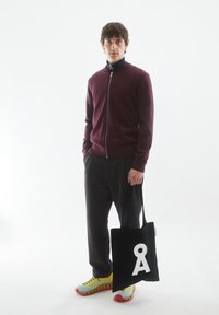 Pull zippé bordeaux, sac fourre-tout noir avec un logo blanc, pantalons en corduroy gris, et baskets multicolores avec des accents jaunes, bleus et rouges.