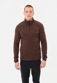 Pullover in pile marrone con colletto a zip a un quarto, tessuto strutturato, maniche lunghe e logo nero sul petto. Indossato con pantaloni scuri.