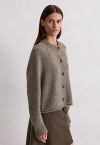 Frau mit langen braunen Haaren, die eine gesprenkelte graue Strickjacke mit Knöpfen und einen dunkeloliven Rock trägt, vor einer schlichten weißen Wand.