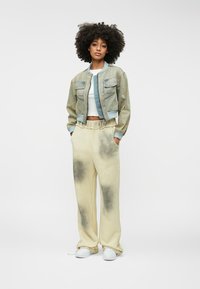 Svijetloplava traper jakna bomber s dva džepa na prsima, u kombinaciji s krem hlačama od dukserice koje imaju izbljedjeli crni uzorak. Model nosi bijele tenisice.