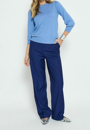 Un pull bleu clair avec un col et des poignets côtelés, associé à un pantalon large à rayures navy. Bracelets en argent et chaussures ornées.