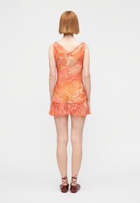 Γυναίκα με κοντά κόκκινα μαλλιά που φοράει αμάνικο διαφανές πορτοκαλί μίνι φόρεμα με σχέδια και καφέ δερμάτινα παπούτσια με κορδόνια, στεκόμενη με την πλάτη προς το μέρος μας.