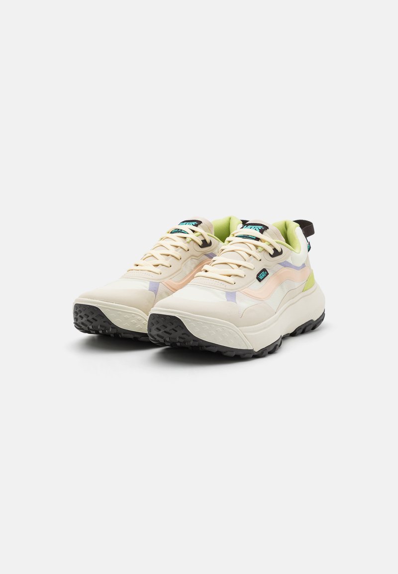 Zapatillas beige con una suela de goma texturizada, detalles en púrpura y verde, y una lengua y cuello acolchados. Presenta cordones y un tirador.