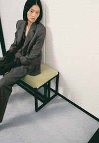 Traje gris texturizado con solapas anchas, sentado en un banco de marco metálico negro con un cojín verde claro, frente a un fondo blanco.