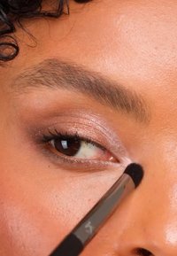 Nærbilde av et øye med myk, glitrende øyenskygge i rosa og brune toner, påført med en liten børste på glatt, teksturert hud.