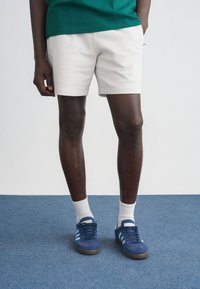 Calções brancos feitos de tecido macio, combinados com ténis azuis com riscas azul claro, meias brancas até ao meio da canela, em pé sobre um tapete azul.