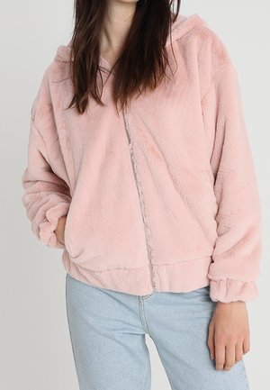 Kvinde iført en lys pink, blød hættetrøje med lynlås og hætte, kombineret med lyseblå jeans, stående med den ene hånd i lommen.