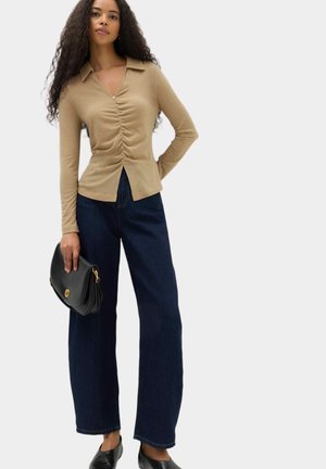 Kvinde med langt krøllet hår iført beige ruchet top, mørke vidde jeans, sorte sko og holder en sort håndtaske.