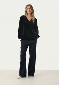 Zwarte fluwelen langemouwen top met een V-hals, gecombineerd met marineblauwe wijde broek en zilveren flats, op een neutrale achtergrond.