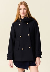 Cappotto in lana nero doppiopetto con bottoni dorati, due tasche frontali e colletto alto. Texture liscia e silhouette strutturata.