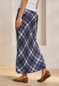 Jupe maxi en tartan marine avec des motifs en damier blancs et bleu clair, fabriquée en tissu lisse, associée à des chaussures plates marron.