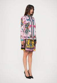 Vrouw in een roze blouse met knopen, bedrukt met vlinders en kroonmotieven, gecombineerd met een geplooide, meerkleurige bloemenrok en zwarte hakken.