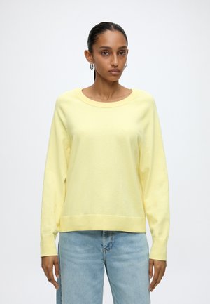 Femme debout, de face, portant un pull jaune pâle à manches longues et un jean bleu clair, sur fond blanc uni.