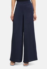 Granatowe spodnie o szerokim kroju, wykonane z gładkiego, lekkiego materiału, z wysokim stanem i zwiewnym fasonem, zestawione z jasnoniebieskimi sandałami na obcasie.