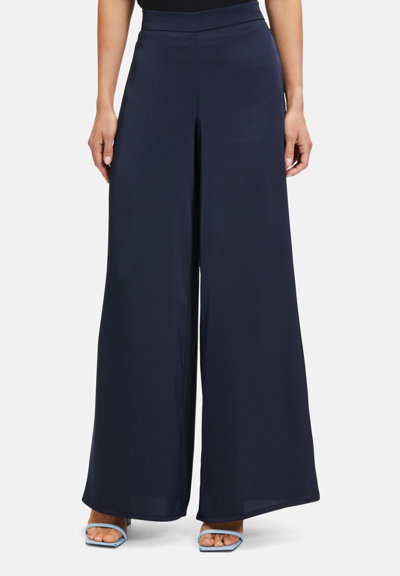 Granatowe spodnie o szerokim kroju, wykonane z gładkiego, lekkiego materiału, z wysokim stanem i zwiewnym fasonem, zestawione z jasnoniebieskimi sandałami na obcasie.