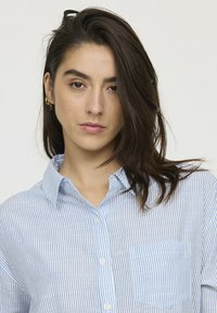 Chemise à rayures bleu et blanche à boutons, avec col, poche avant et texture de tissu lisse. Le mannequin porte de petites boucles d'oreilles en or.