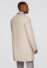 Cappotto beige con colletto, dal taglio dritto e con dettagli minimi. Texture liscia, design sino al ginocchio, tasche laterali visibili.