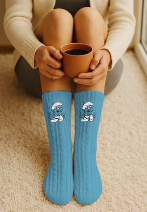 Person sitzt im Schneidersitz auf dem Teppich und hält eine braune Tasse, trägt blaue, kabelgestrickte Socken mit einer gehenden blauen Cartoon-Figur.