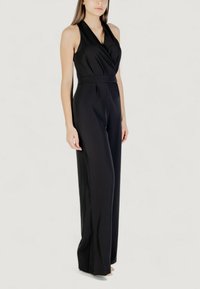 Donna che indossa una tuta nera senza maniche con colletto a V avvolgente e pantaloni a gamba larga, in piedi contro uno sfondo chiaro e uniforme.