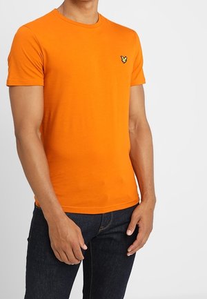 T-shirt en coton orange à manches courtes et col rond. Présente un petit logo jaune sur la poitrine gauche. Porté avec un jean bleu foncé.