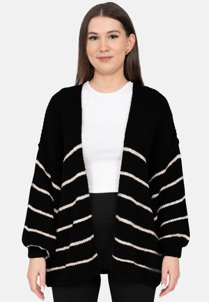 Jeune femme aux cheveux bruns raides portant des boucles d'oreilles en or, un haut blanc, un pantalon noir et un cardigan noir à rayures horizontales blanches.