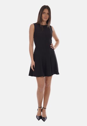 Donna in piedi, che indossa un vestito nero senza maniche con una gonna a campana e tacchi neri con cinturino alla caviglia su uno sfondo bianco semplice.