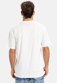 Camiseta blanca de manga corta con un corte relajado, textura suave, cuello redondo y una pequeña etiqueta de logo verde en el dobladillo, mostrada desde la parte posterior.